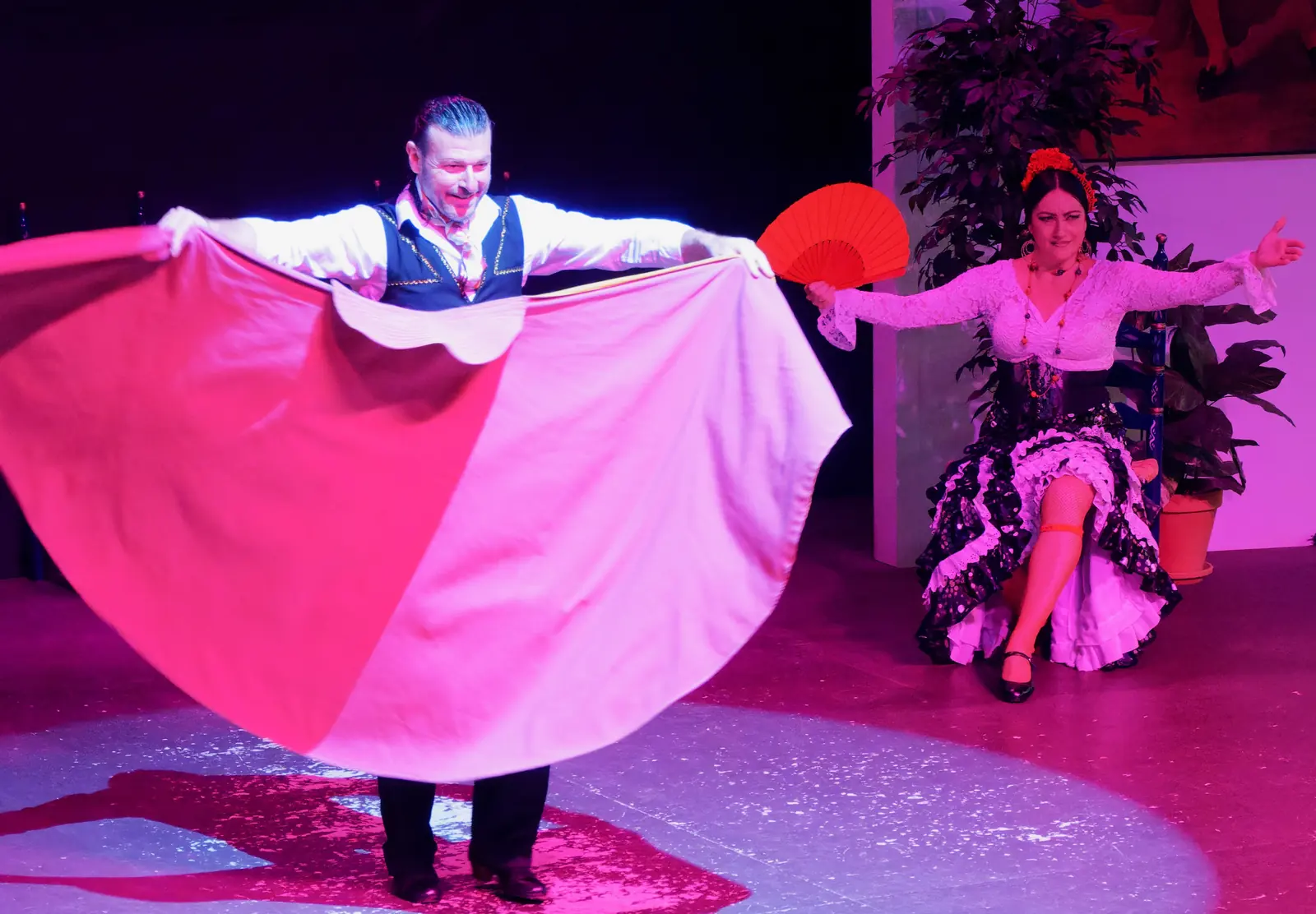 Un ballerino maschio tende un ampio mantello rosa e arancione accanto a una ballerina seduta con un ventaglio rosso