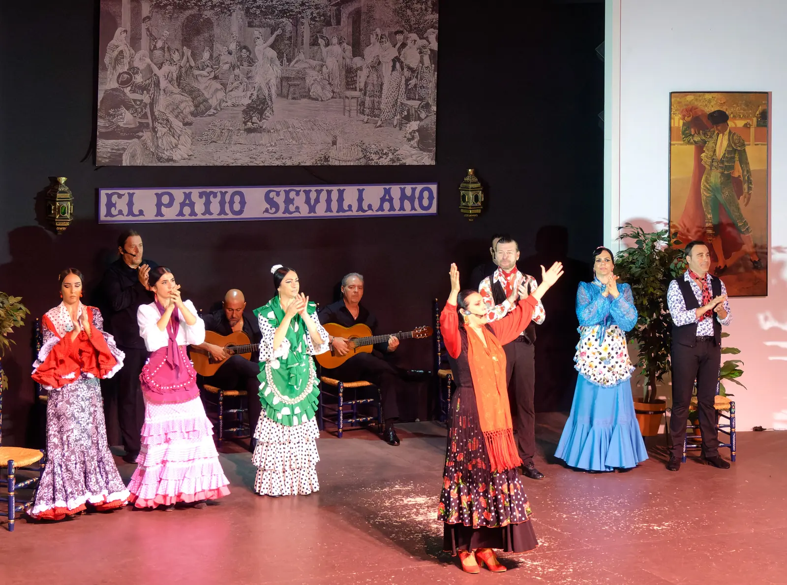 L'intera compagnia in piedi sul palco mentre applaude, ballerine in abiti a pois e con volant e due chitarristi seduti dietro, sotto un'insegna in piastrelle blu con la scritta El Patio Sevillano