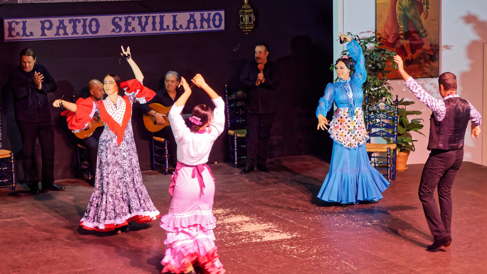 Flamenkové tanečnice v červených, růžových a modrých volánových šatech zvedají kastaněty na jevišti zalitém červeným světlem, za nimi sedí kytaristé a modrý kachlový nápis hlásá El Patio Sevillano
