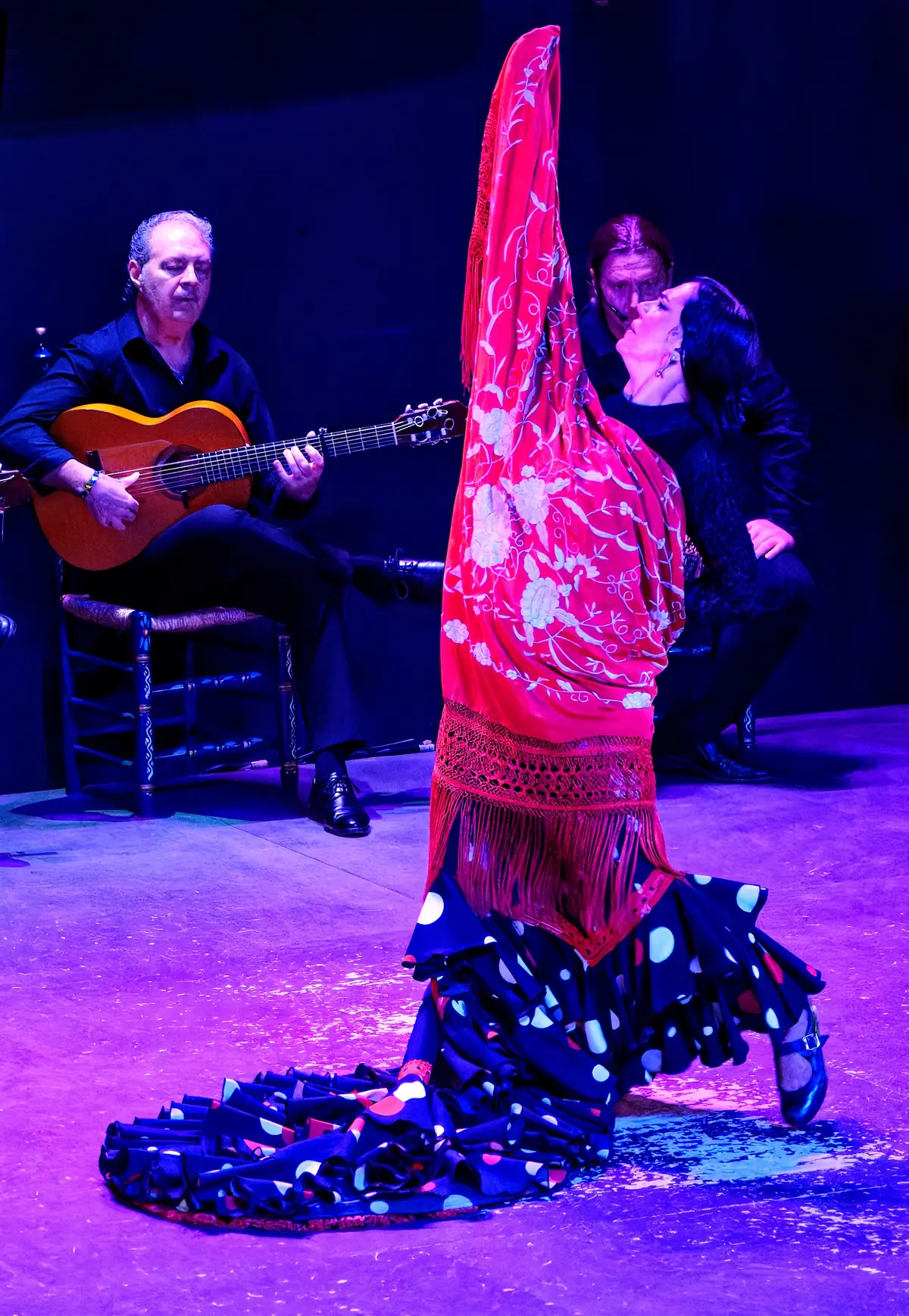 Una ballerina in abito nero a pois con uno scialle rosso drappeggiato dalle braccia sollevate
