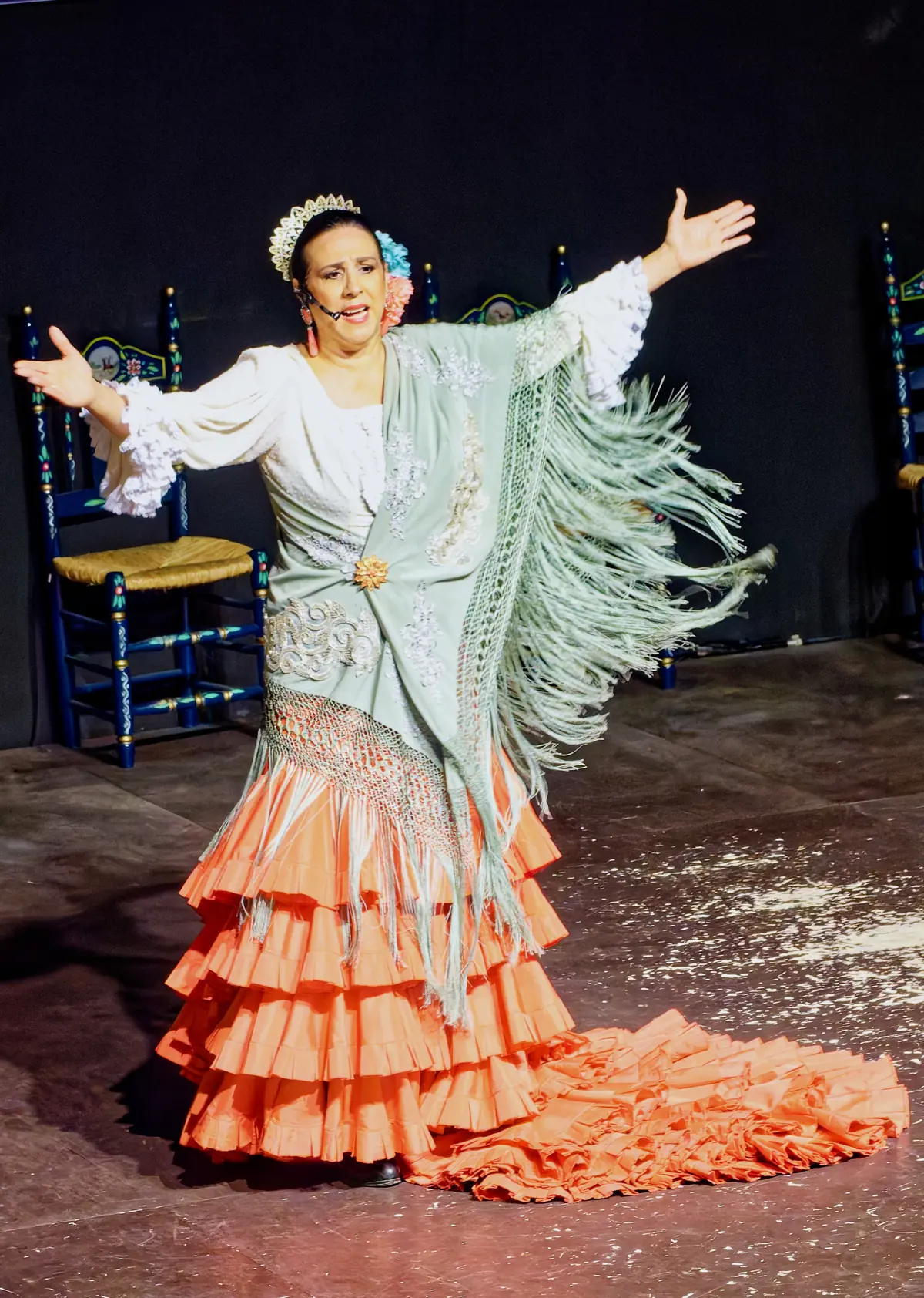 Una ballerina in un abito color pesca con volant e uno scialle leggero verde pallido con frange, le braccia spalancate su un palco buio